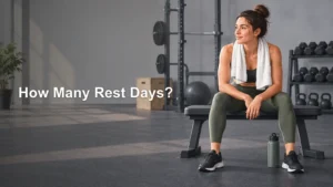 A woman in workout clothes sits on a gym bench with a towel around her neck, surrounded by weights and a water bottle; text reads “How Many Rest Days?”